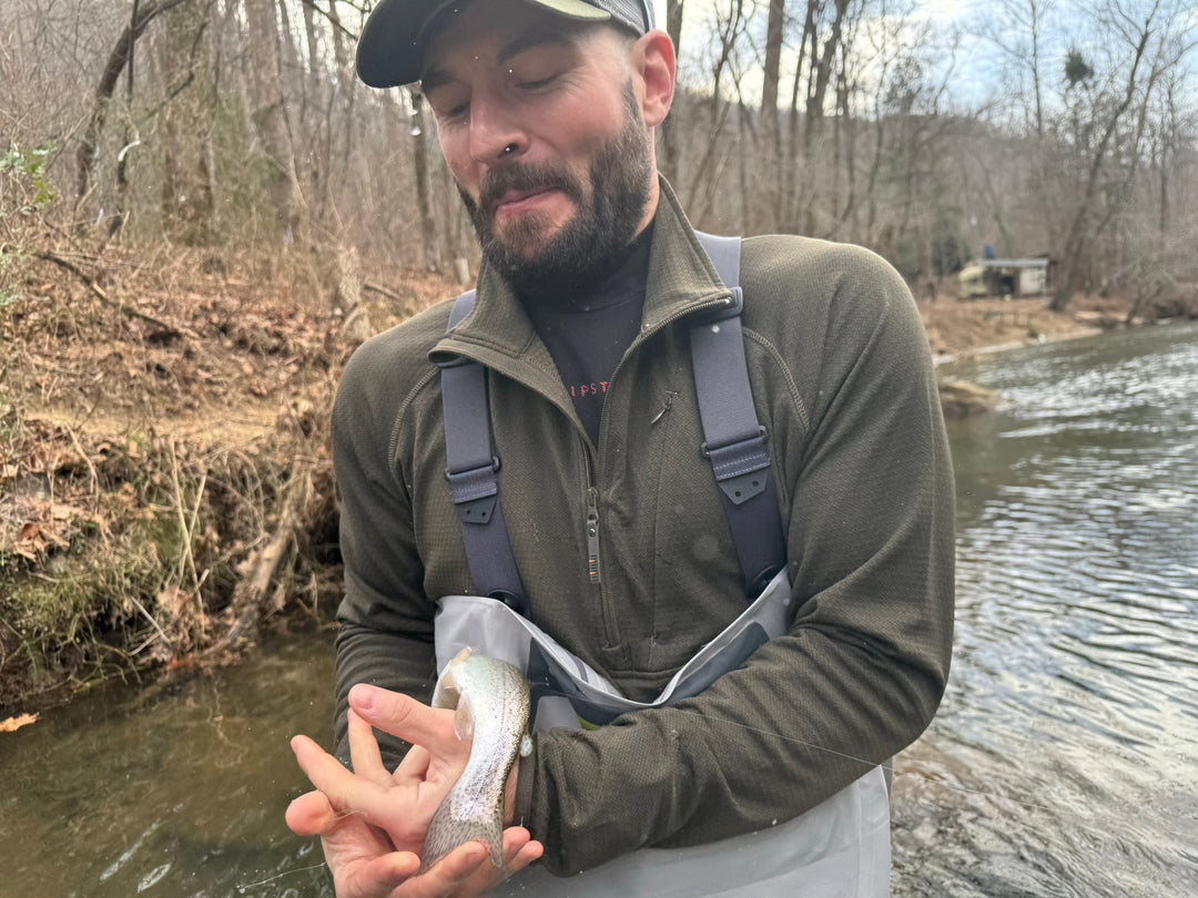 A client with a beard and hat starts laughing while standing in the stream struggles while fumbling around trying to hold his first slippery trout on the fly he caught. Winter scene on the Green River in Saluda, NC.