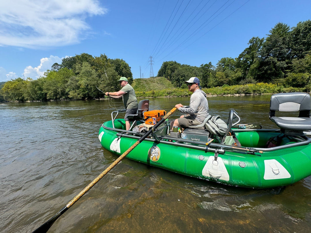 Guide Caleb Simmons rows a green Sotar raft with a client along a stretch of the French Broad River in search of trophy smallmouth bass. There are green trees and bluebird skies on an exceptionally hot day in Asheville, NC.